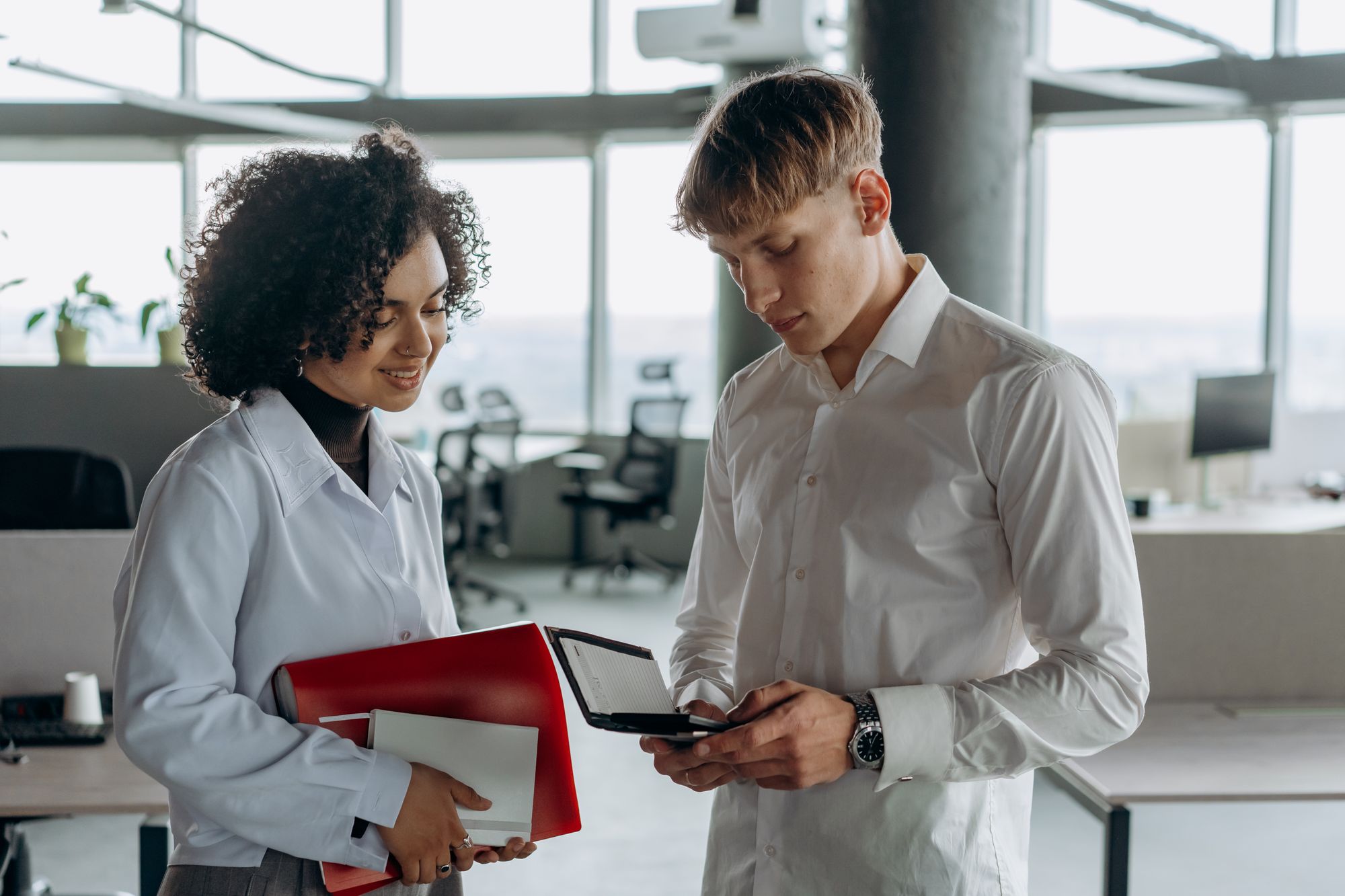 Two colleagues review figures with a calculator and folders in a bright open office.