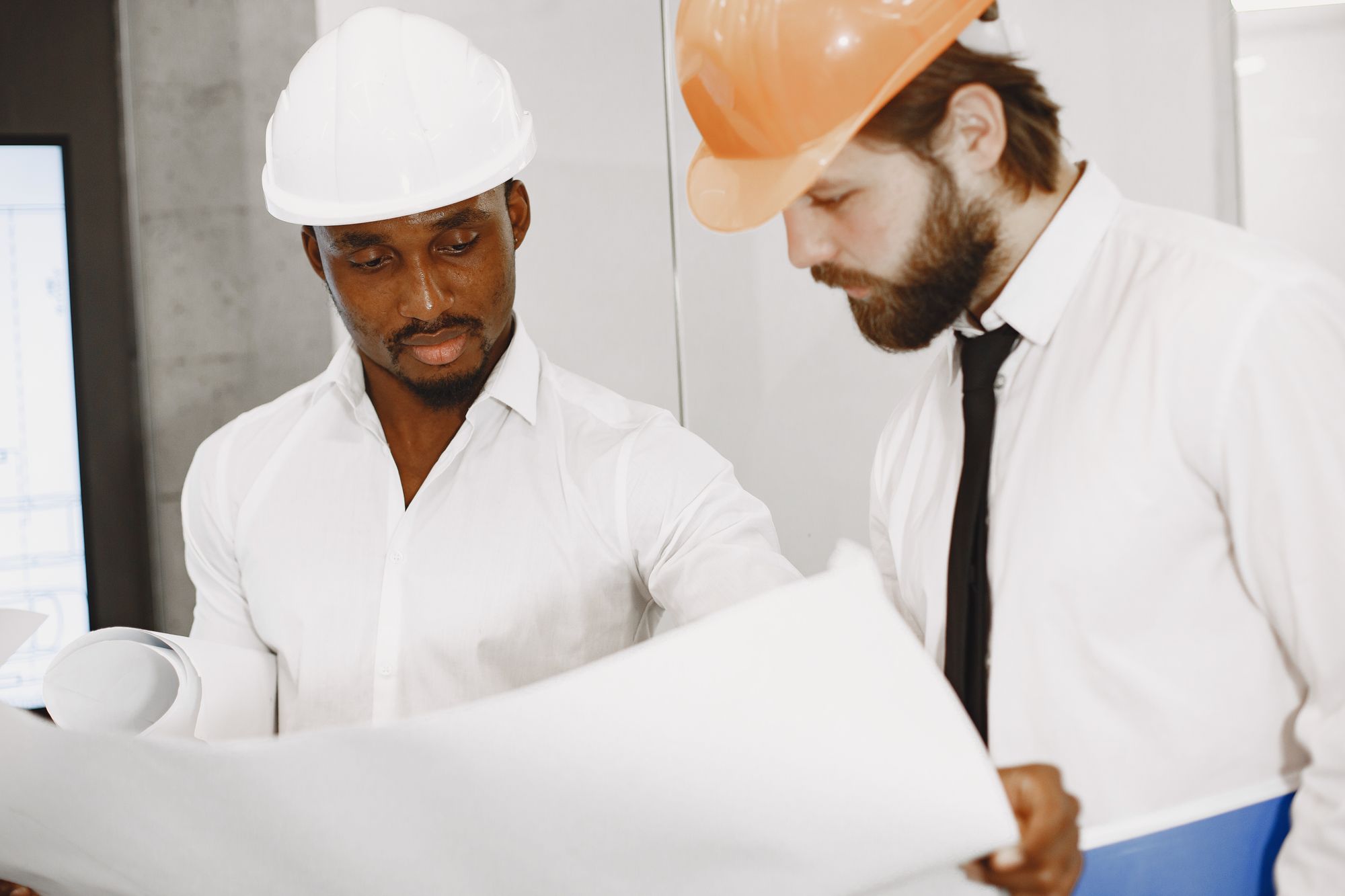 Two construction professionals wearing hard hats review a large set of plans indoors.
